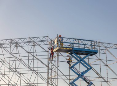 Bir binayı onarmak için mavi bir vinç kullanılıyor. İki adam vinç üzerinde çalışıyor, biri sarı bir miğfer takıyor.