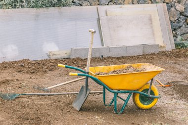 Üzerinde kürek olan sarı bir el arabası. Kürek yerde, el arabasının yanında.