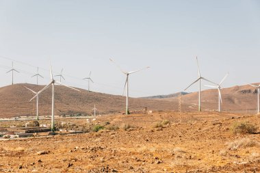 Bir grup rüzgar türbini bir çöldeki yamaçta. Yel değirmenleri uzundur ve yayılır, bazıları kameraya daha yakın, bazıları ise daha uzaktadır. Sonsuzluk ve güç kavramı