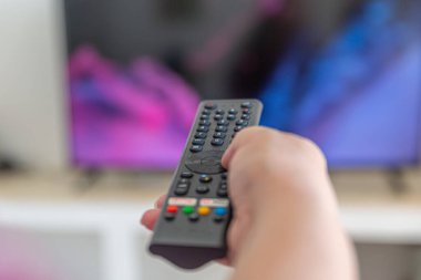 Bir kişi televizyonun önünde uzaktan kumanda tutuyor. Uzaktan kumandanın siyah bir rengi var ve kişinin elinde.