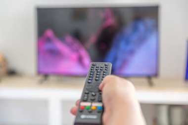 Arka planda renkli ama bulanık bir ekran olan bir televizyonu çalıştırmak için uzaktan kumanda kullanan birinin yakın çekimi. Konforlu ev ortamı.