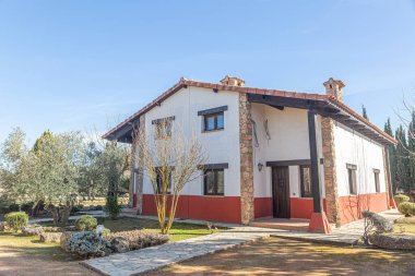 Kırmızı çizgili büyük beyaz bir ev çimenli bir bahçede oturur. Evin dış yüzeyi taştan, dış yüzeyi de kırmızı. Bahçe ağaçlar ve çalılarla dolu ve eve giden bir yol var.