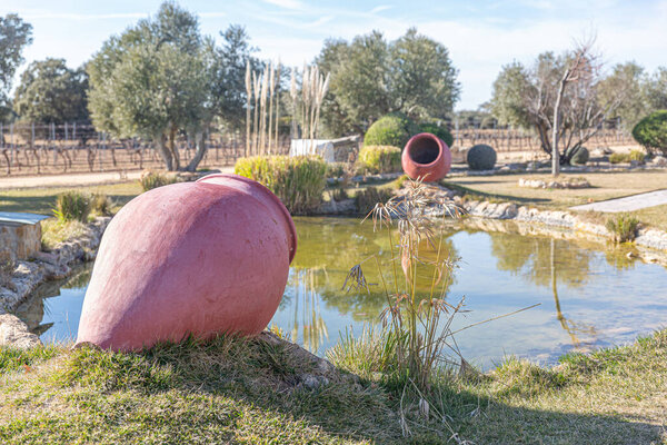 A large red vase sits on the grass next to a pond. The vase is surrounded by plants and trees, creating a peaceful and serene atmosphere