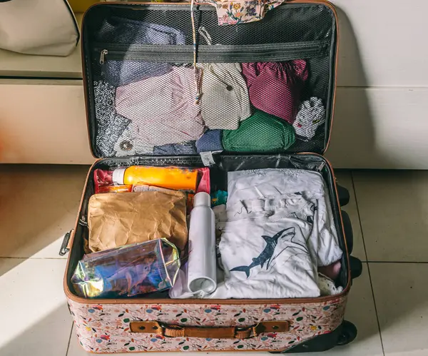 Bir bavul açık ve giysilerle dolu. Yerde küçük bavulun yanında bir şişe güneş kremi var.