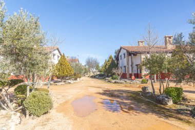Toprak yolu ve birkaç evi olan küçük bir kasaba. Evler beyaz ve kırmızı. Yol çamurlu ve su birikintisi var. Gökyüzü mavi ve berrak