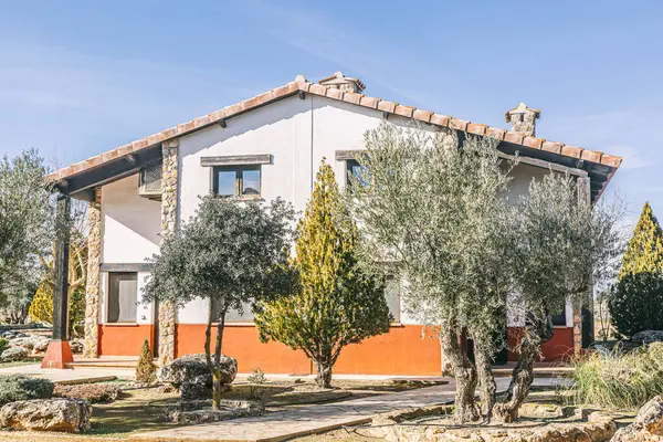 Kırmızı tuğladan duvarı ve kırmızı çatısı olan büyük beyaz bir ev. Evin önünde büyük bir ağaç ve arkasında daha küçük bir ağaç var. Evin etrafı bahçeyle çevrili ve yolu ona çıkıyor.