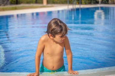 Küçük bir çocuk havuzda duruyor, suya bakıyor. Yeşil bir mayo giyiyor.