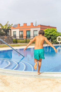 Bir çocuk havuzda oynuyor. Havuz mavi ve beyaz bir merdiveni var. Çocuk yeşil bir mayo giyiyor.