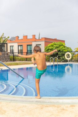 Genç bir çocuk havuzun kenarında duruyor, tek ayak üzerinde dengede duruyor. Havuz bir çitle çevrili ve arka planda bir bina var. Çocuk eğleniyor gibi görünüyor.