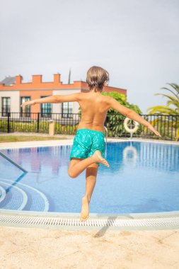 Mavi mayo giymiş bir çocuk havuza atlıyor. Havuz çitlerle çevrili ve nöbetçi bir cankurtaran var.
