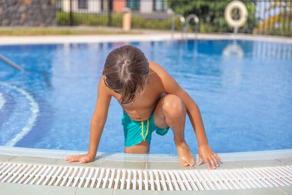 Мальчик стоит на краю бассейна и смотрит на воду. На нём зелёный купальник и мокрые волосы. Сцена игривая и светлая