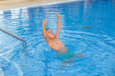 Genç bir çocuk suda, su sıçratıyor ve oynuyor. Yeşil bir gömlek ve şort giyiyor.