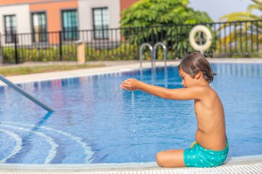 Küçük bir çocuk havuzda oturup suyla oynuyor. Havuz çitlerle çevrili ve yakınlarda bir cankurtaran standı var. Çocuk eğleniyor gibi görünüyor.