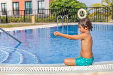 Küçük bir çocuk havuzda oturuyor, ellerini suyun içinde tutuyor. Sahne huzurlu ve rahatlatıcı, çocuk suda eğleniyor.