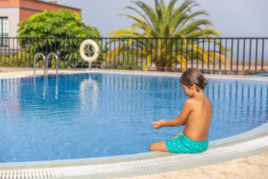 Küçük bir çocuk havuzda oturuyor, suya bakıyor. Havuz bir çitle çevrili ve yakınında bir can yeleği var. Çocuk havuzda eğleniyor gibi görünüyor.