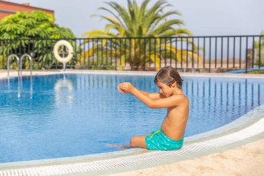 Küçük bir çocuk havuzda oturuyor, ellerini birarada tutuyor. Havuz çitlerle çevrili ve görev başında bir cankurtaran var. Çocuk suda eğleniyor gibi görünüyor.