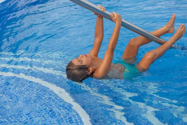 Sudaki bir havuz direğine asılmış genç bir çocuk var. Çocuk mavi bir mayo giyiyor ve eğleniyor.