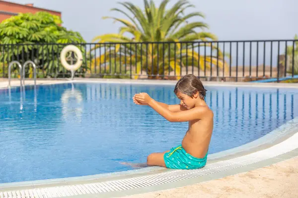 Küçük bir çocuk havuzda oturuyor, ellerini birarada tutuyor. Havuz çitlerle çevrili ve görev başında bir cankurtaran var. Çocuk suda eğleniyor gibi görünüyor.