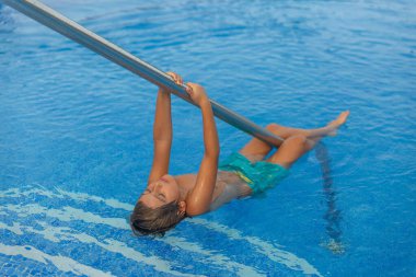 Küçük bir çocuk havuzda metal bir direğe asılıyor. Mavi bir mayo giyiyor ve eğleniyor.
