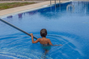Bir çocuk havuzda elinde erişteyle bekliyor. Havuz mavi ve su sakin.