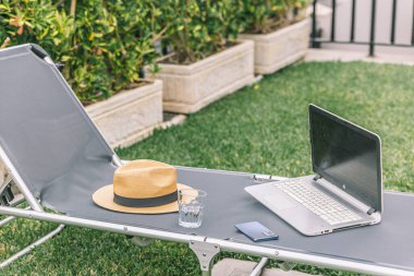 Dizüstü bilgisayar katlanır sandalyede bir şapka ve bir bardak suyun yanında. Sahne, sakin ve sakin bir atmosferi işaret ediyor.