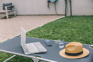 Dizüstü bilgisayar, şezlongda, şapkası ve bir bardak suyla birlikte. Sahne, muhtemelen bir günlük çalışma ya da dışarıda çalışma için sakin ve sakin bir atmosfer öneriyor.