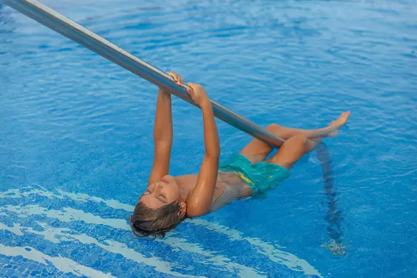 Küçük bir çocuk havuzda metal bir direğe asılıyor. Mavi bir mayo giyiyor ve eğleniyor.