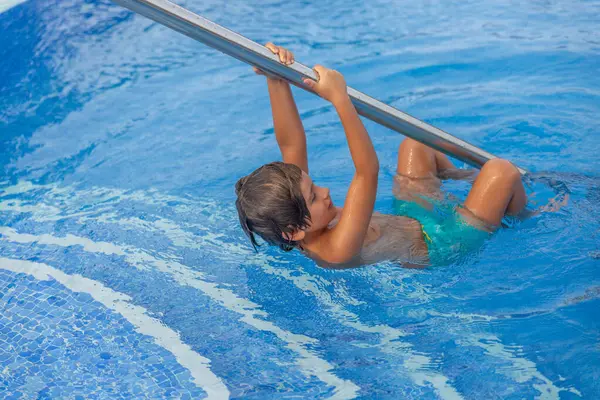 Bir çocuk havuz merdiveninden suya sarkıyor. Yeşil bir mayo giyiyor.
