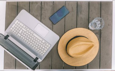 Bir dizüstü bilgisayar, cep telefonu ve şapka ahşap bir masada. Laptop açık ve üstünde de cep telefonu var. Sahne, rahat ve rahat bir atmosferi işaret ediyor.