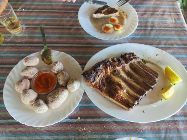 Bir tabak kaburga bir tabak patates ve bir tabak yumurtanın yanındaki masaya konur. Masa renkli bir masa örtüsüyle kaplıdır ve yakınlarda çatal ve bıçak gibi çatallar vardır.