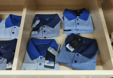 Bir mağazada bir sıra gömlek sergileniyor, bazılarının üzerinde etiket var. Gömlekler mavi ve beyaz polo gömlekler de dahil olmak üzere çeşitli renk ve stillerde.