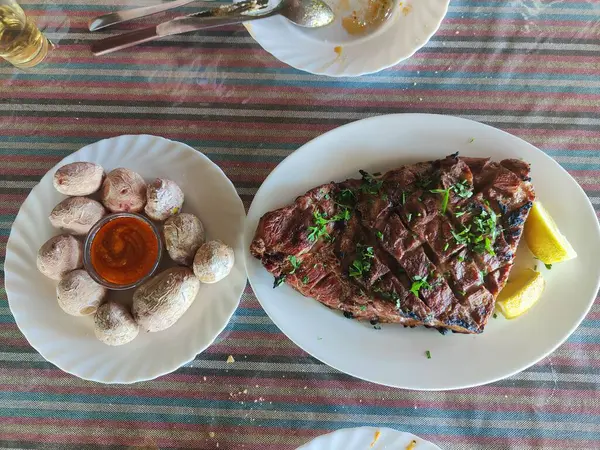 Üzerinde limon dilimleri ve maydanoz olan bir tabak et masanın üzerinde duruyor. Masa kırmızı ve mavi çizgili bir bezle kaplıdır. Masada başka yemek tabakları da var.