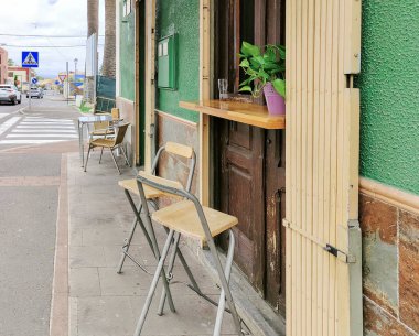 Yeşil binalı, beyaz ve mavi tabelası olan bir sokak manzarası. Bir restoranın dışında birkaç sandalye ve masa var.
