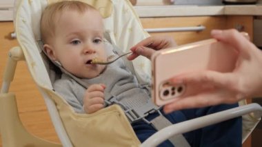 Little baby boy watching video on smartphone during breakfast in highchair. Concept of parenting, healthy nutrition and baby feeding