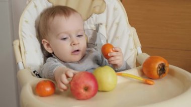 Portrait of little baby boy sitting in highchair and eating fruits at kitchen. Concept of parenting, healthy nutrition and baby feeding