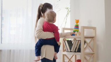 Mutlu gülümseyen anne oturma odasındaki raftan küçük oğluna oyuncak veriyor. Bebek gelişimi, aile oyunları, ilk adımlar, ebeveynlik ve bakım.