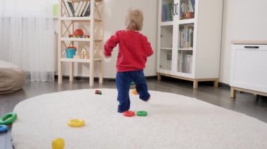Küçük çocuk halıda yürümeyi ve renkli oyuncaklar toplamayı öğreniyor. Bebek gelişimi, aile oyunları, ilk adımlar, ebeveynlik ve bakım.