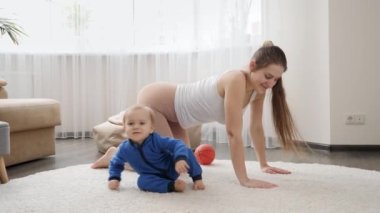 Güzel, genç bir anne, oturma odasında halının üzerinde oturan küçük oğlunun yanında spor yapıyor. Aile sağlığı, aktif yaşam tarzı, ebeveynlik ve çocuk gelişimi.
