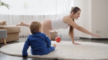 Tatlı erkek bebek yerde oturup, evde yoga ve spor yapan annesine bakıyor. Aile sağlığı, aktif yaşam tarzı, ebeveynlik ve çocuk gelişimi
