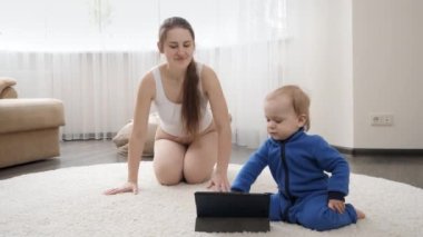 Küçük oğlu olan genç bir anne halı üzerinde spor yapıyor ve tablet bilgisayarda eğitim videosu izliyor. Aile sağlığı, aktif yaşam tarzı, ebeveynlik ve çocuk gelişimi.