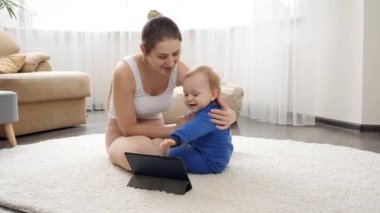 Gülümseyen genç anne tablet bilgisayarda video izlerken bebeğine masaj yapıyor ve vücudunu esnetiyor. Aile sağlığı, aktif yaşam tarzı, ebeveynlik ve çocuk gelişimi