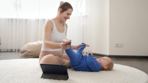 年轻母亲在家里观看网上视频课程 并在地毯上伸展她的男婴身体 家庭保健 积极的生活方式 养育子女和儿童发展 — 图库视频影像