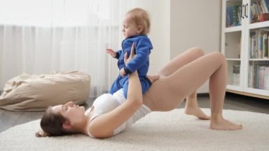 Genç, sevgi dolu bir anne, evde spor yaparken bebeğini kucağında tutuyor. Aile sağlığı, aktif yaşam tarzı, ebeveynlik ve çocuk gelişimi.