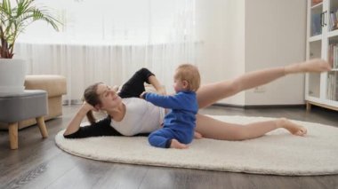 Sevimli erkek bebek yerde yatan ve esneme egzersizleri yapan genç anneyi izliyor. Aile sağlığı, aktif yaşam tarzı, ebeveynlik ve çocuk gelişimi.