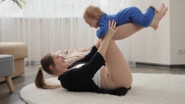 Mutlu gülümseyen anne, evde yoga ve spor yaparken bebeğini kucağına alıp sallıyor. Aile sağlığı, aktif yaşam tarzı, ebeveynlik ve çocuk gelişimi.