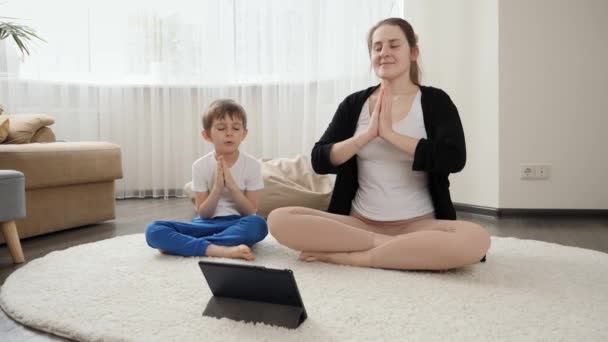 在平板电脑上的在线课程中 一个小男孩 他的妈妈在一边呼吸 一边坐在瑜伽里 家庭保健 积极的生活方式 养育子女和儿童发展 — 图库视频影像