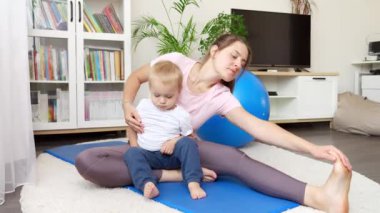 Güzel genç bir kadın ve küçük bir oğlu var spor paspası üzerinde oturuyor ve bacaklarını esnetiyor. Evde sağlık, spor ve yoga kavramı