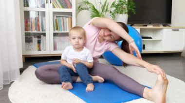 Beden eğitimi paspasında oturan ve spor antrenmanından önce bacaklarını esneten annesine bakan tatlı bir çocuk. Evde sağlık, spor ve yoga kavramı