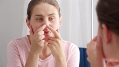 Genç bir kadın aynaya bakıyor ve burnunda sivilceler çıkıyor. Kadın yaşlanma, cilt sorunları, yüz bakımı ve bakımı kavramı