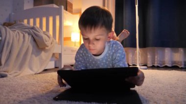 Yatak odasındaki yumuşak halıda yatarken pijamalı çocuk tablet bilgisayarda oynuyor. Çocukların eğitimi, gelişimi, alet kullanan çocuklar gizlilik, mahremiyet.